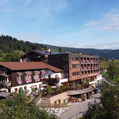 A large hotel building in a rural setting with green hills in the background. The sky is clear and the landscape looks inviting. | © Das Alphotel Hirschegg Hotel GmbH | Mark Kerwell