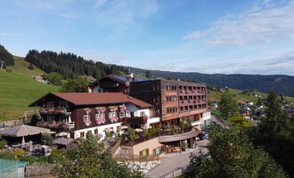 Ein großes Hotelgebäude in ländlicher Umgebung mit grünen Hügeln im Hintergrund. Der Himmel ist klar und die Landschaft wirkt einladend. | © Das Alphotel Hirschegg Hotel GmbH | Mark Kerwell