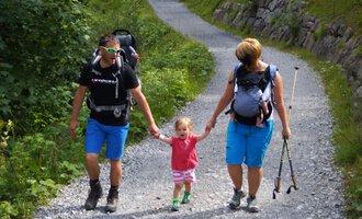 A family is hiking on a narrow path through nature. The parents are holding their small child's hands and carrying backpacks. | © Austrian Ski- & Service Ranch | Trixi Leitner