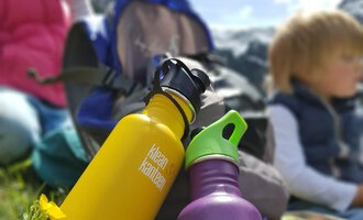 Zwei bunte Wasserflaschen in Gelb und Lila stehen im Vordergrund auf einer grünen Wiese. Im Hintergrund sind Kinder und ein Rucksack zu sehen, mit Bergen und blauem Himmel. | © Austrian Ski- & Service Ranch | Trixi Leitner