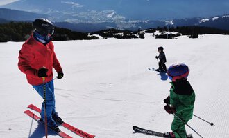 Zwei Kinder stehen auf Skiern in einem sonnigen Skigebiet. Im Hintergrund sind schneebedeckte Berge und eine klare Landschaft zu sehen. | © Austrian Ski- & Service Ranch | Trixi Leitner