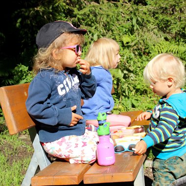 Drei Kinder sitzen auf einer Bank im Freien. Sie genießen Snacks und spielen mit Spielzeug auf einem Tisch. | © Kleinwalsertal Tourismus | Britta Maier
