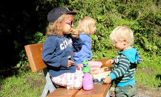 Drei Kinder sitzen auf einer Bank im Freien. Sie genießen Snacks und spielen mit Spielzeug auf einem Tisch. | © Kleinwalsertal Tourismus | Britta Maier