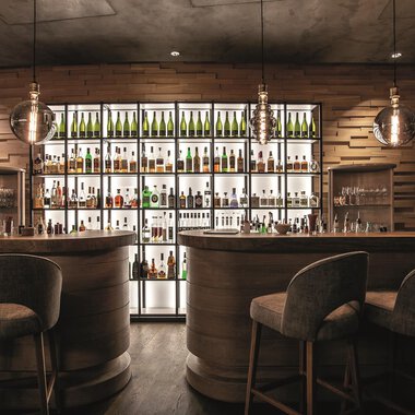 Eine stilvolle Bar mit Holzinterieur und einem beleuchteten Regal voller Flaschen. Die Sitzgelegenheiten sind gemütlich und modern. | © A-ROSA Ifen Hotel Kleinwalsertal