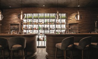 Eine stilvolle Bar mit Holzinterieur und einem beleuchteten Regal voller Flaschen. Die Sitzgelegenheiten sind gemütlich und modern. | © A-ROSA Ifen Hotel Kleinwalsertal