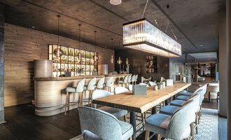 Ein eleganter Innenraum eines modernen Restaurants mit einer langen Holztafel und stilvollen Stühlen. Im Hintergrund befindet sich eine gut beleuchtete Bar mit einer Vielzahl von Getränken. | © A-ROSA Ifen Hotel Kleinwalsertal