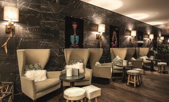Ein modernes Café mit gemütlichen Sitzecken und stilvollem Dekor. Die Wände sind mit kunstvollen Bildern dekoriert und es gibt sanfte Beleuchtung. | © A-ROSA Ifen Hotel Kleinwalsertal