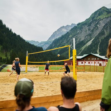A beach volleyball game in the mountains, surrounded by a picturesque landscape. The players are actively on the sand court while spectators watch. | © Kleinwalsertal Tourismus | Oliver Farys