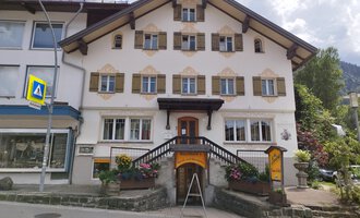 A charming, traditional building with many windows and a large entrance. The house has an appealing facade with colorful flowers at the entrance. | © Kleinwalsertal Tourismus | N. Lughammer