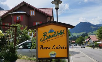 A rustic sign from "Bergblick" with the inscription "Bura Lädele". In the background, typical Alpine houses and mountains can be seen. | © Kleinwalsertal Tourismus | N. Lughammer