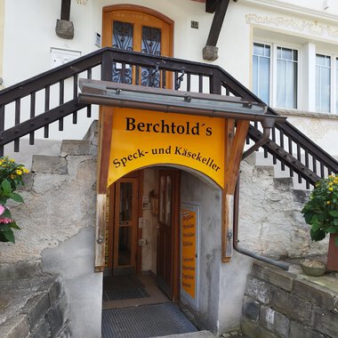 Ein Eingangsbereich mit einer farbenfrohen Beschilderung für Berchtold's Speck- und Käsekeller. Treppen führen in den Laden, umgeben von Blumen in Töpfen. | © Kleinwalsertal Tourismus | N. Lughammer