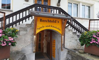 Ein Eingangsbereich mit einer farbenfrohen Beschilderung für Berchtold's Speck- und Käsekeller. Treppen führen in den Laden, umgeben von Blumen in Töpfen. | © Kleinwalsertal Tourismus | N. Lughammer