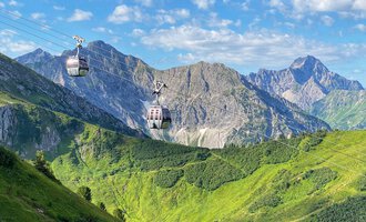 A cable car glides over green hills and mountains. The sky is clear and blue, with a few clouds. | © OBERSTDORF · KLEINWALSERTAL BERGBAHNEN