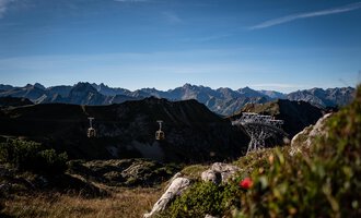 Eine Seilbahn fährt über eine grüne Wiese mit Felsen. Im Hintergrund sind majestätische Berge und ein klarer Himmel zu sehen. | © OBERSTDORF · KLEINWALSERTAL BERGBAHNEN