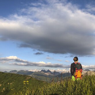 Eine Person steht auf einer Wiese und blickt auf die Berge. Der Himmel ist blau mit einigen Wolken. | © Kleinwalsertal Tourismus | Louisa Hieke