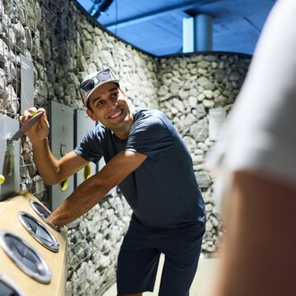 Ein Mann lächelt und interagiert mit einer Spiel- oder Installationswand. Die Umgebung besteht aus Steinen und modernem Design. | © Kleinwalsertal Tourismus |  Oliver Farys