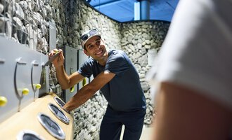 Ein Mann lächelt und interagiert mit einer Spiel- oder Installationswand. Die Umgebung besteht aus Steinen und modernem Design. | © Kleinwalsertal Tourismus |  Oliver Farys