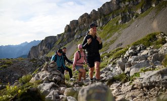 A group of hikers is climbing a rocky path in the mountains. The landscape is green and characterized by impressive rock formations. | © Bergschule Kleinwalsertal @Bergschulel Oliver Farys
