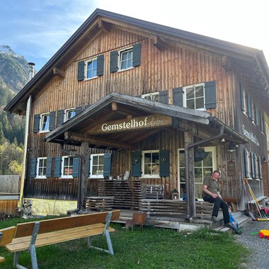 A traditional wooden house named "Gemstelhof" stands in a rural setting. Benches are placed in front of the building, and a person is visible, while the mountains are seen in the background. | © Bernhards Gemstel Hofladen | Bernhard Heim