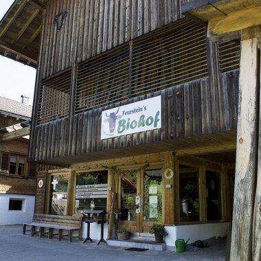 Ein Holzgebäude mit einem Schild, das „BioHof“ anzeigt. Daneben stehen Tische und Stühle im Außenbereich, umgeben von weiteren traditionellen Gebäuden. | © Kleinwalsertal Tourismus