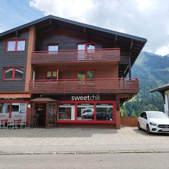 Ein modernes Gebäude mit zwei Etagen in den Bergen. Das Erdgeschoss beherbergt das Restaurant "sweetchill" mit großen Fenstern. | © Kleinwalsertal Tourismus | N. Lughammer