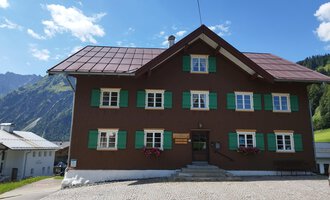 A traditional old house with a brown facade and green shutters. In the background, mountains and a blue sky are visible. | © Kleinwalsertal Tourismus | Nicole Lughammer