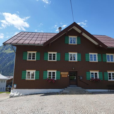 A traditional old house with a brown facade and green shutters. In the background, mountains and a blue sky are visible. | © Kleinwalsertal Tourismus | Nicole Lughammer