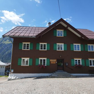 Ein traditionelles altes Haus mit brauner Fassade und grünen Fensterläden. Im Hintergrund sind Berge und ein blauer Himmel sichtbar. | © Kleinwalsertal Tourismus | Nicole Lughammer