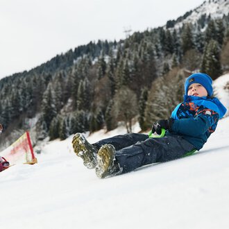A sled lies in the snow, surrounded by a wintry mountain landscape. The sun shines out from behind the mountains. | © Kleinwalsertal Tourismus | Oliver Farys