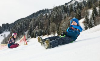 A sled lies in the snow, surrounded by a wintry mountain landscape. The sun shines out from behind the mountains. | © Kleinwalsertal Tourismus | Oliver Farys