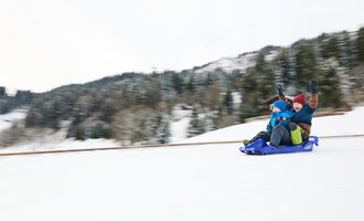 Ein Schlitten liegt im Schnee, umgeben von Bergen. Die Sonne strahlt im Hintergrund und schafft eine winterliche Atmosphäre. | © Kleinwalsertal Tourismus | Oliver Farys