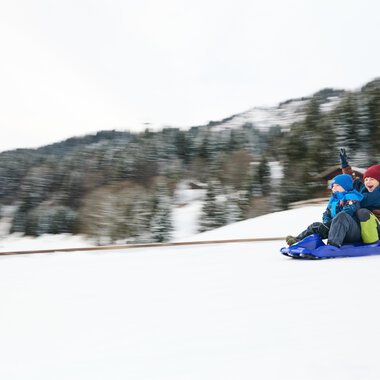 Ein Schlitten liegt im Schnee, umgeben von Bergen. Die Sonne strahlt im Hintergrund und schafft eine winterliche Atmosphäre. | © Kleinwalsertal Tourismus | Oliver Farys