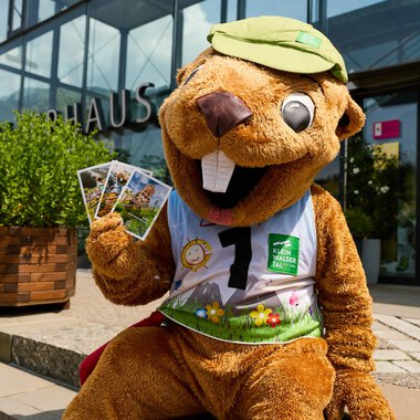 Ein lustiger Wasservogel-Maskottchen sitzt vor einem Gebäude und hält einige Postkarten in der Hand. Es trägt ein grünes Cap und ein buntes Outfit. | © Kleinwalsertal Tourismus | Oliver Farys