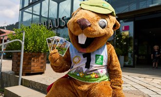 A funny water bird mascot sits in front of a building and holds some postcards in its hand. It is wearing a green cap and a colorful outfit. | © Kleinwalsertal Tourismus | Oliver Farys