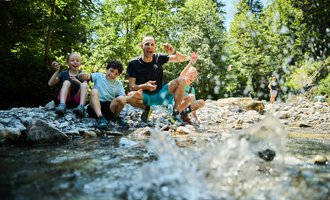 Eine Gruppe von Kindern sitzt am Ufer eines glasklaren Baches und hat Spaß. Im Hintergrund sind grüne Bäume und eine sommerliche Landschaft zu sehen. | © Kleinwalsertal Tourismus | Oliver Farys