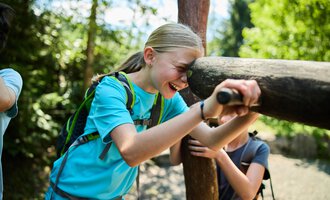 Ein fröhliches Kind, das lachend an einem Holzpfahl lehnt, umgeben von einem bewaldeten Bereich. Die Sonne scheint durch die Bäume und schafft eine freundliche Atmosphäre. | © Kleinwalsertal Tourismus | Oliver Farys