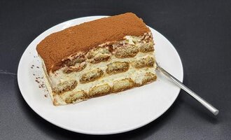 Ein Stück Tiramisu auf einem weißen Teller. Das Dessert ist mit Kakao bestäubt und hat eine schichtweise Struktur.