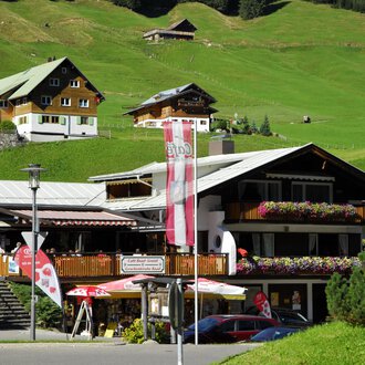 Ein charmantes Gasthaus mit blühenden Balkonen in einer grünen Berglandschaft. Im Hintergrund sind weitere traditionelle Hütten zu sehen. | © Cafe Baad Grund | Roman Schuster