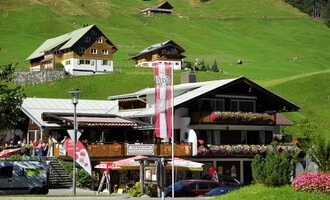 Ein charmantes Gasthaus mit blühenden Balkonen in einer grünen Berglandschaft. Im Hintergrund sind weitere traditionelle Hütten zu sehen. | © Cafe Baad Grund | Roman Schuster