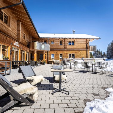 A cozy wooden building in winter with a terrace.  
Chairs and tables are on the snow-covered ground, surrounded by snow-covered trees.