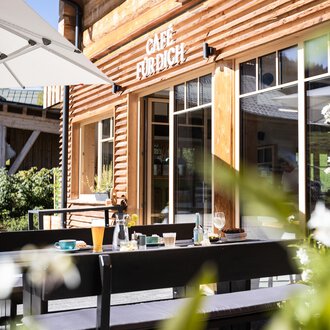 Ein einladendes Café mit Holzfassade und einem sonnigen Außenbereich. Tische sind bereit für Gäste, um die frische Luft zu genießen.