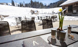 Ein Tisch im Freien mit einer Speisekarte und einer kleinen Blume. Im Hintergrund sind schneebedeckte Berge und eine gemütliche Atmosphäre zu sehen.