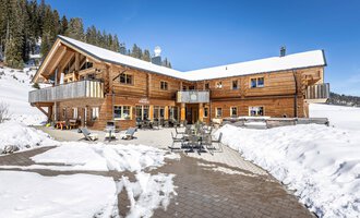 Ein gemütliches Holzhaus im Schnee mit einer Terrasse und gemütlichen Sitzgelegenheiten. Im Hintergrund sind verschneite Bäume und ein klarer blauer Himmel zu sehen.