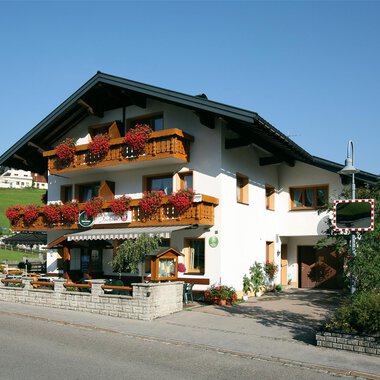 Ein malerisches Gebäude im alpinen Stil mit blühenden Balkonen. Es liegt in einer ruhigen, grünen Umgebung. | © Cafe Jochum | Bernadette Reinmuth