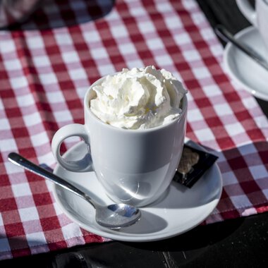 Eine Tasse mit Sahne und Löffel steht auf einem Tisch mit rot-weißem kariertem Tischdecke. Im Hintergrund sind weitere Tassen zu sehen. | © Kleinwalsertal Tourismus | Dominik Berchtold