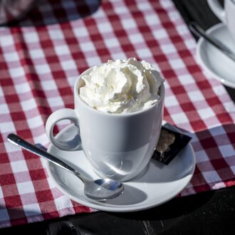Eine Tasse mit Sahne und Löffel steht auf einem Tisch mit rot-weißem kariertem Tischdecke. Im Hintergrund sind weitere Tassen zu sehen. | © Kleinwalsertal Tourismus | Dominik Berchtold