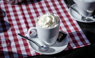 Eine Tasse mit Sahne und Löffel steht auf einem Tisch mit rot-weißem kariertem Tischdecke. Im Hintergrund sind weitere Tassen zu sehen. | © Kleinwalsertal Tourismus | Dominik Berchtold