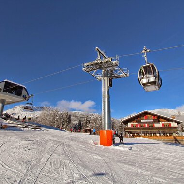 Eine Skiliftstation in einer verschneiten Berglandschaft. Im Hintergrund sind ein Chalet und ein klarer blauer Himmel zu sehen.