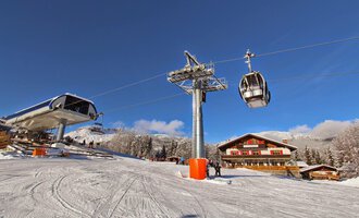 Eine Skiliftstation in einer verschneiten Berglandschaft. Im Hintergrund sind ein Chalet und ein klarer blauer Himmel zu sehen.