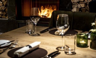 An elegant table with dishes and glasses, decorated for a dinner. In the background, a fireplace is burning, creating a cozy atmosphere. | © A-ROSA Ifen Hotel Kleinwalsertal